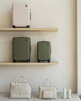 A display of three suitcases on two wooden shelves. The top shelf features a check-in medium in desert taupe and two olive-green suitcases in check-in medium and carry-on sizes, while the bottom shelf shows a metro weekender and metro duffel in ivory (vegan leather) and two kiyo uvc water bottles in castle rock arranged in a minimalist setting.