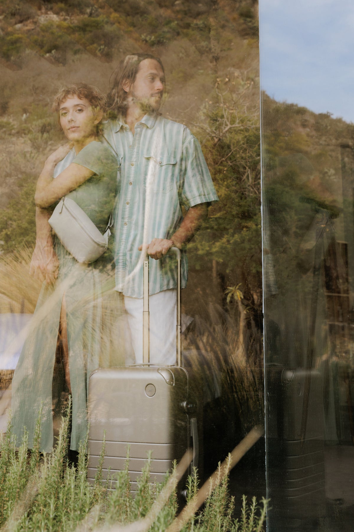 A reflective glass image of a couple standing close together, with the woman wearing a metro sling in ivory (vegan leather) and the man with his hand on a hybrid carry-on in champagne. Behind them, lush outdoor scenery is visible through the glass.
