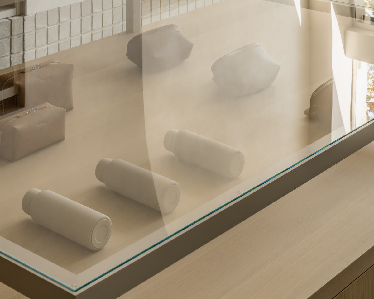 A display case with several kiyo uvc water bottles in castle rock, metro sling, and metro toiletry cases in saddle tan (vegan leather) behind glass. The arrangement is neat, set against a neutral background.