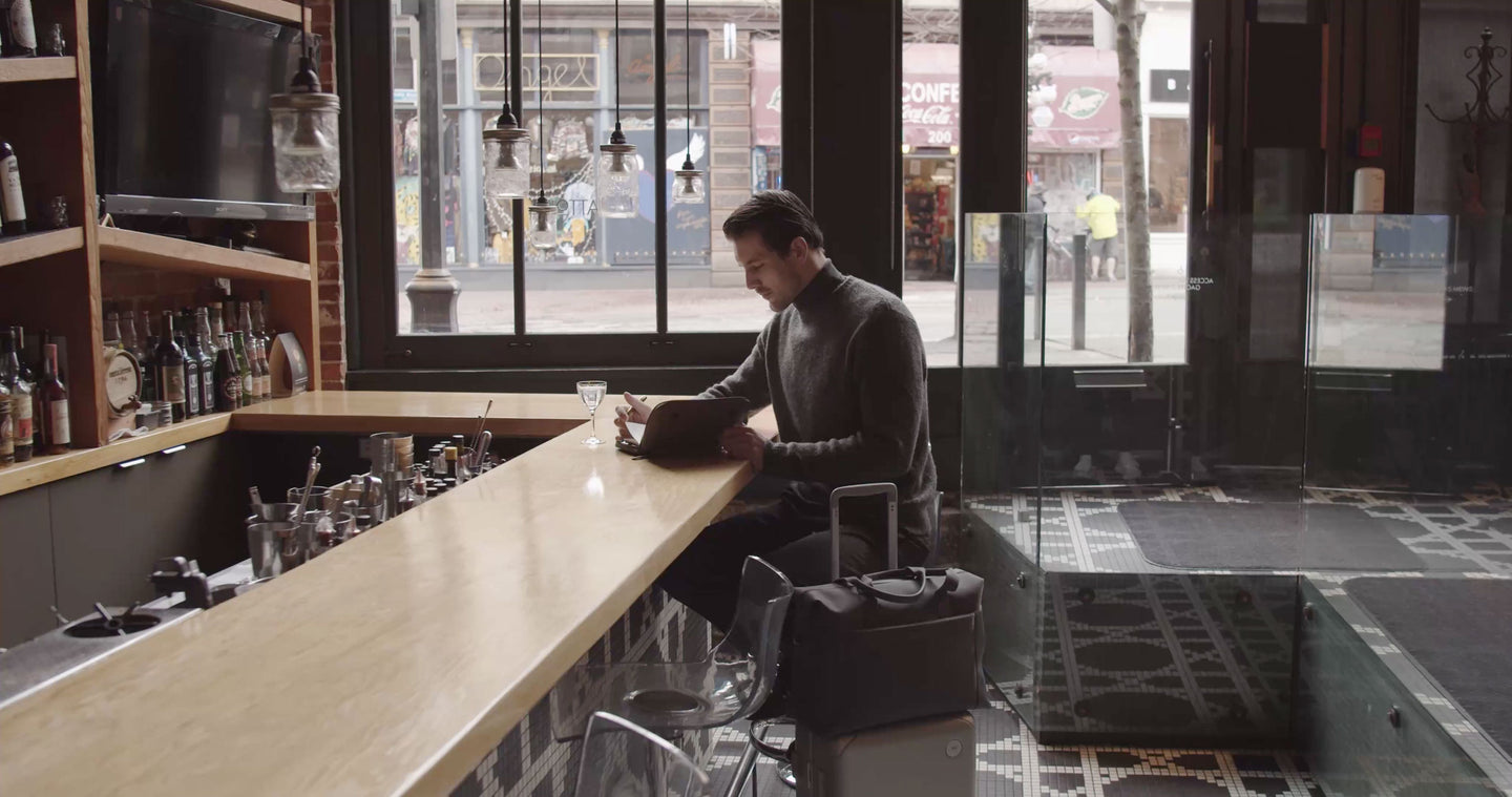 A man sitting in a bar with a Hybrid Carry-On and a Metro Duffel on top of it