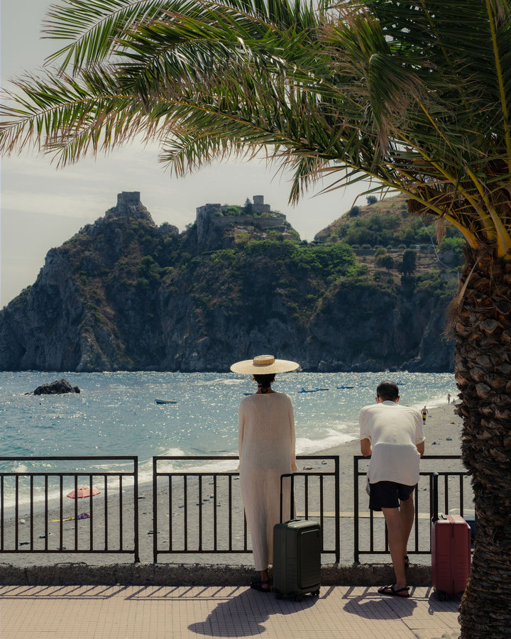 A serene beach scene with two people standing by a railing, gazing out at the ocean and distant rocky cliffs. The woman, in a long light-coloured dress and wide-brimmed hat, stands with a carry-on pro in olive green while the man leans on the railing with a carry-on pro in terracotta beside him. A palm tree frames the scene, adding to the tranquil vacation atmosphere.