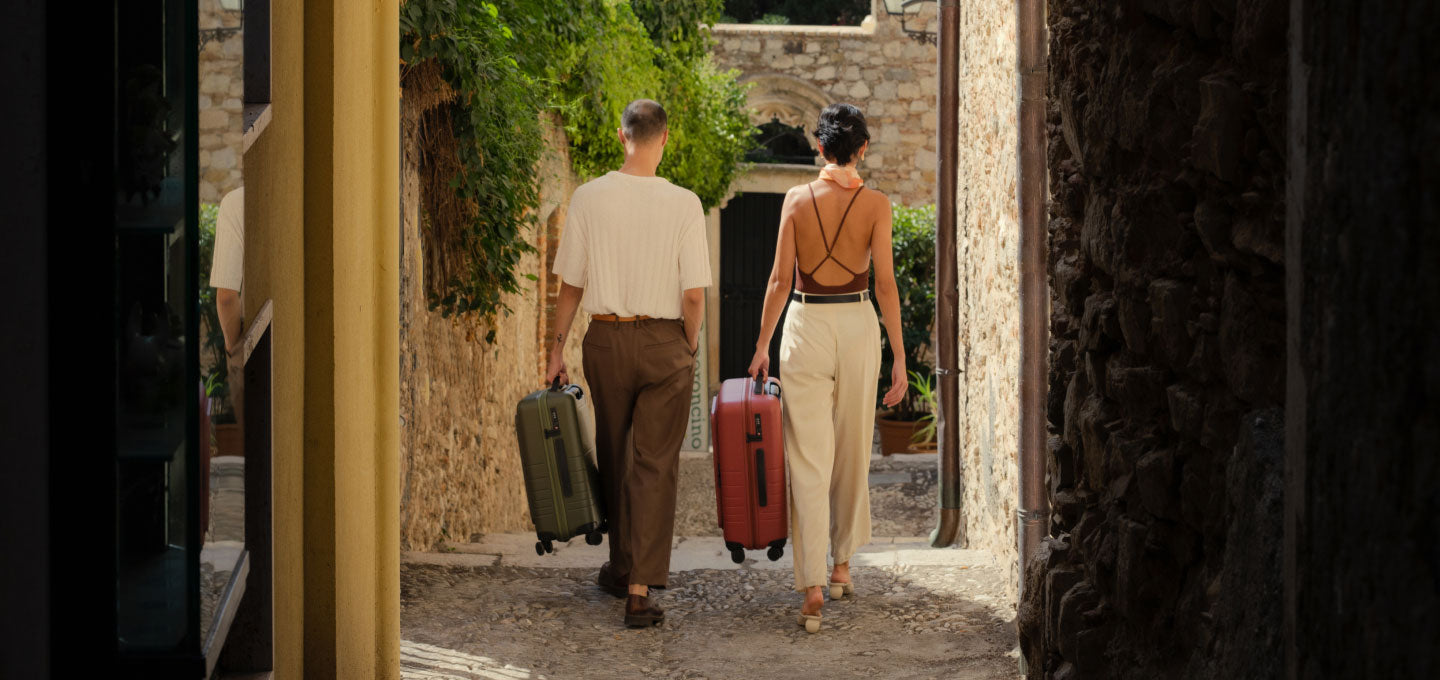 A couple walking down a narrow cobblestone alley lined with stone walls and greenery. The woman, dressed in light trousers and a strappy top, carries a carry-on pro in terracotta, while the man, in brown trousers and a white top, carries a carry-on pro in olive green. They walk side by side toward a quaint, sunlit destination.