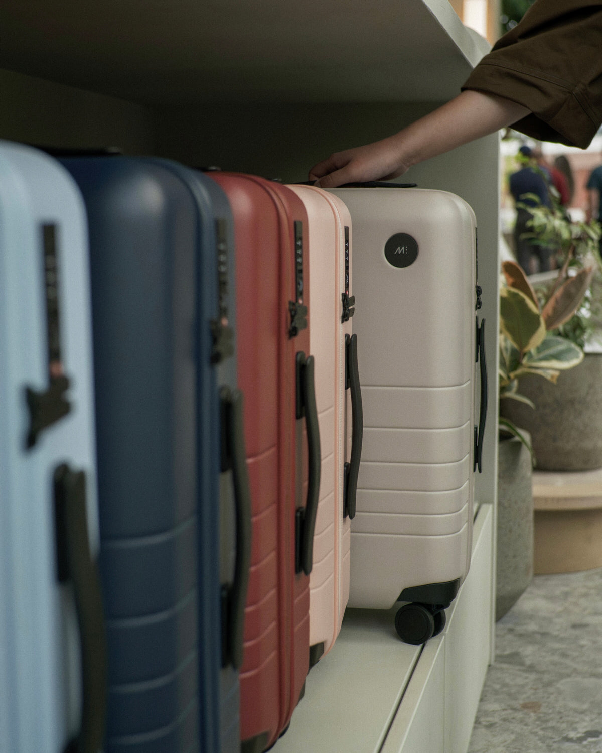 The Classic collection displayed on a shelf with a person's hand resting on a Desert Taupe suitcase.