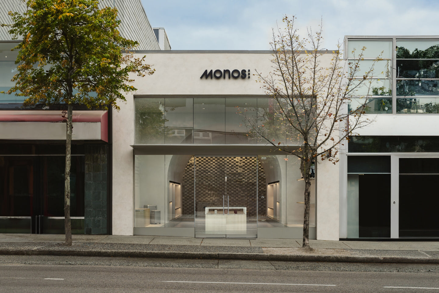 The exterior of a Monos retail store with large glass windows showcasing the inside of the store. The entrance is framed by a simple, modern design, with the Monos logo in black above the door, and trees flanking the storefront.