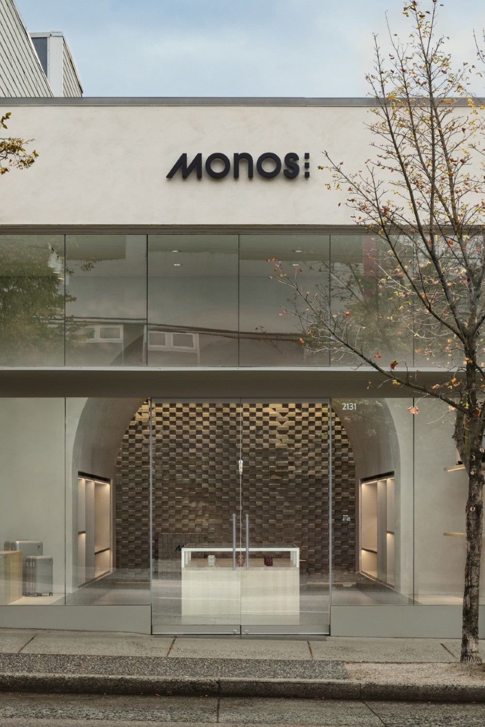 The exterior of a Monos retail store with large glass windows showcasing the inside of the store. The entrance is framed by a simple, modern design, with the Monos logo in black above the door, and a tree flanking the storefront.